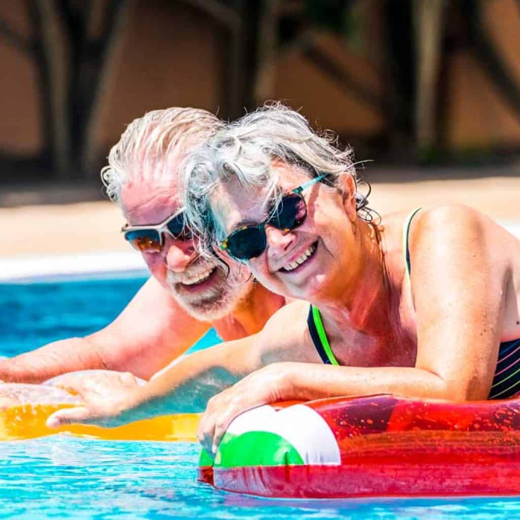 People retired old senior man and woman couple enjoying summer swimming pool leisure activity lay down on coloured trendy fashion lilos inflatable mattress on blue water at home or hotel activity