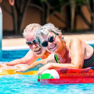 People retired old senior man and woman couple enjoying summer swimming pool leisure activity lay down on coloured trendy fashion lilos inflatable mattress on blue water at home or hotel activity
